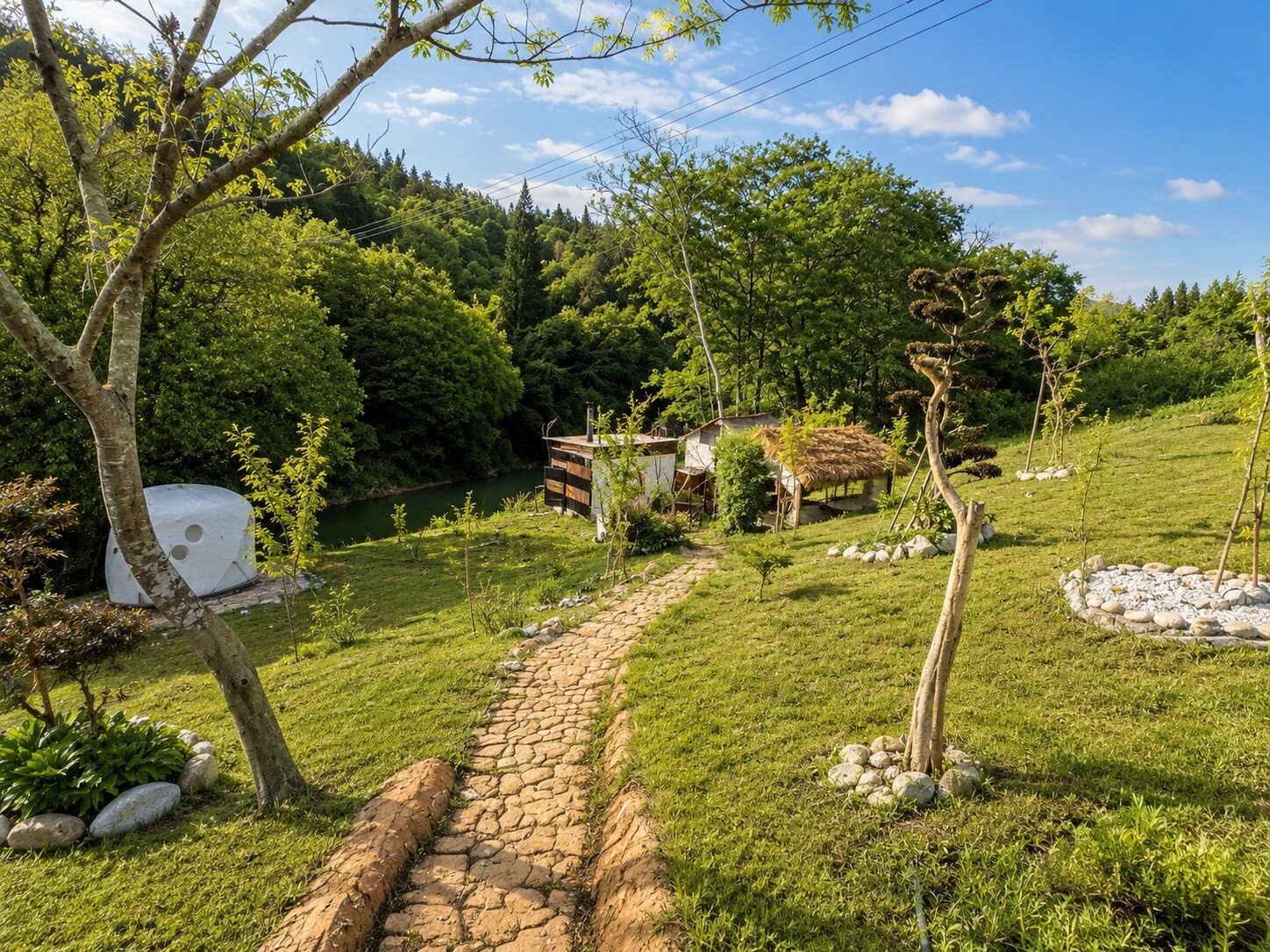 緑の山に囲まれた敷地を抜ける石畳の小道と、川辺に建つ手作りのサウナ小屋